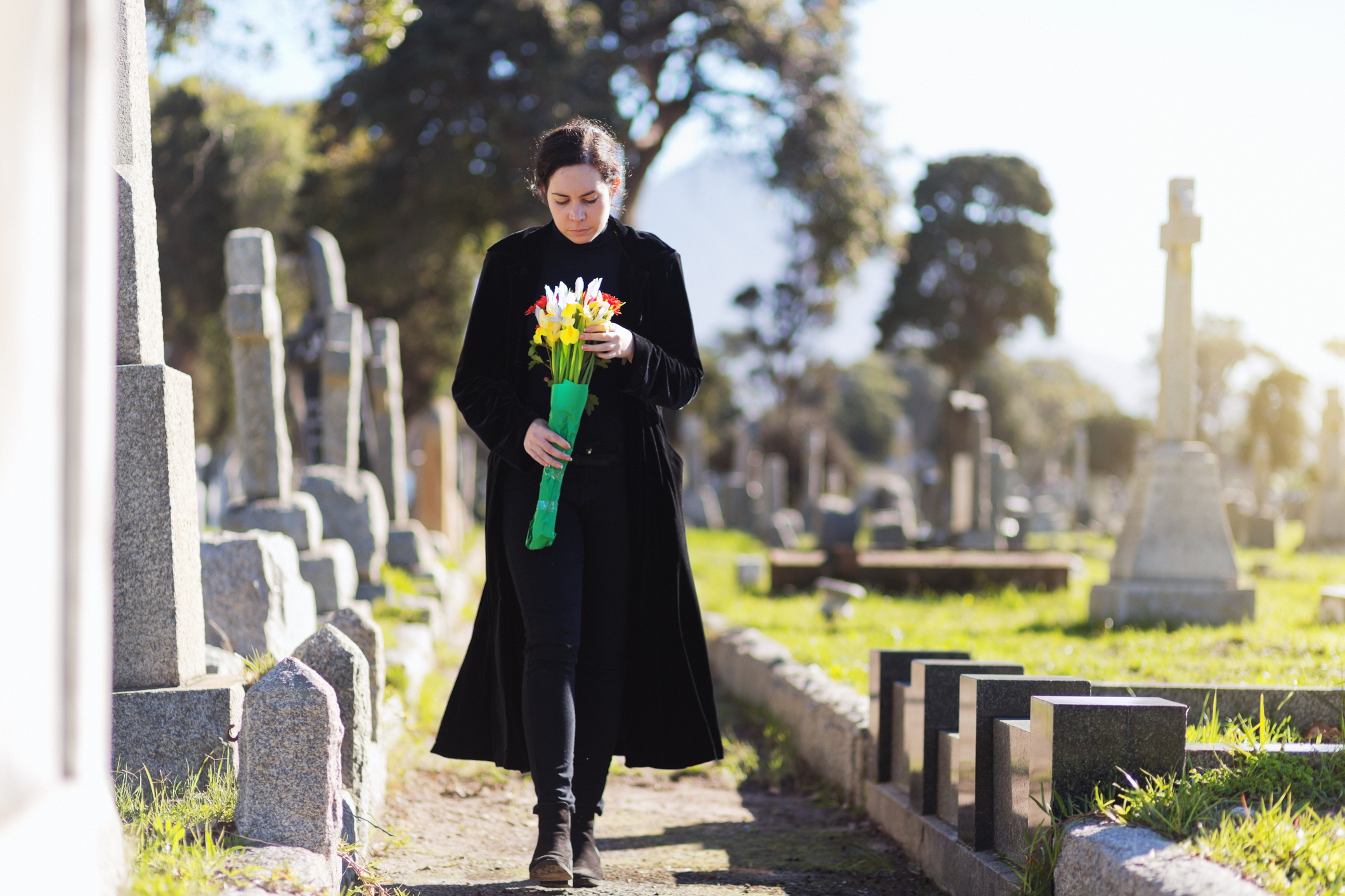 A young woman in black walks through cemetery headstones carrying flowers to the grave of someone she misses
