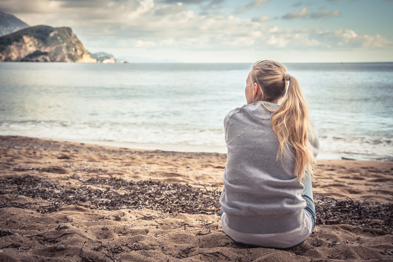 Pensive lonely young woman sitting on beach hugging her knees and looking into the distance with hope