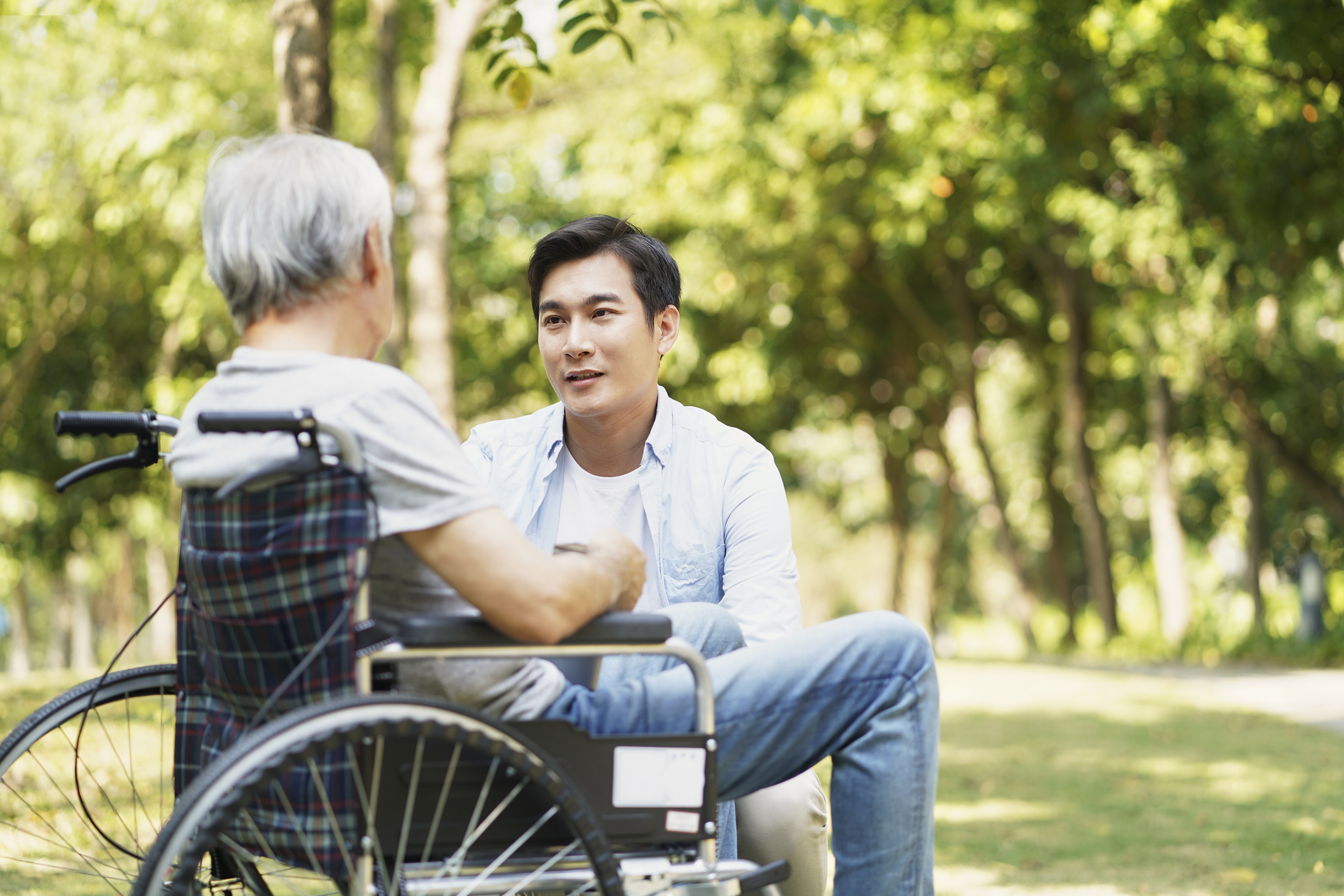 Young asian adult son chatting with wheel chair bound father outdoors in park