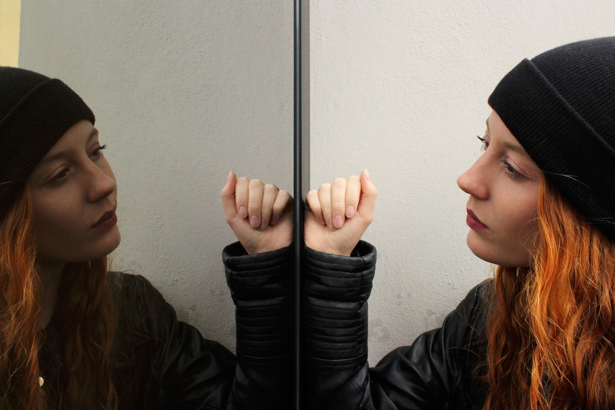 Girl with long red hair wearing a black woolen cap and a black leather jacket looking at herself mirrored on a glass door with a thoughtful expression