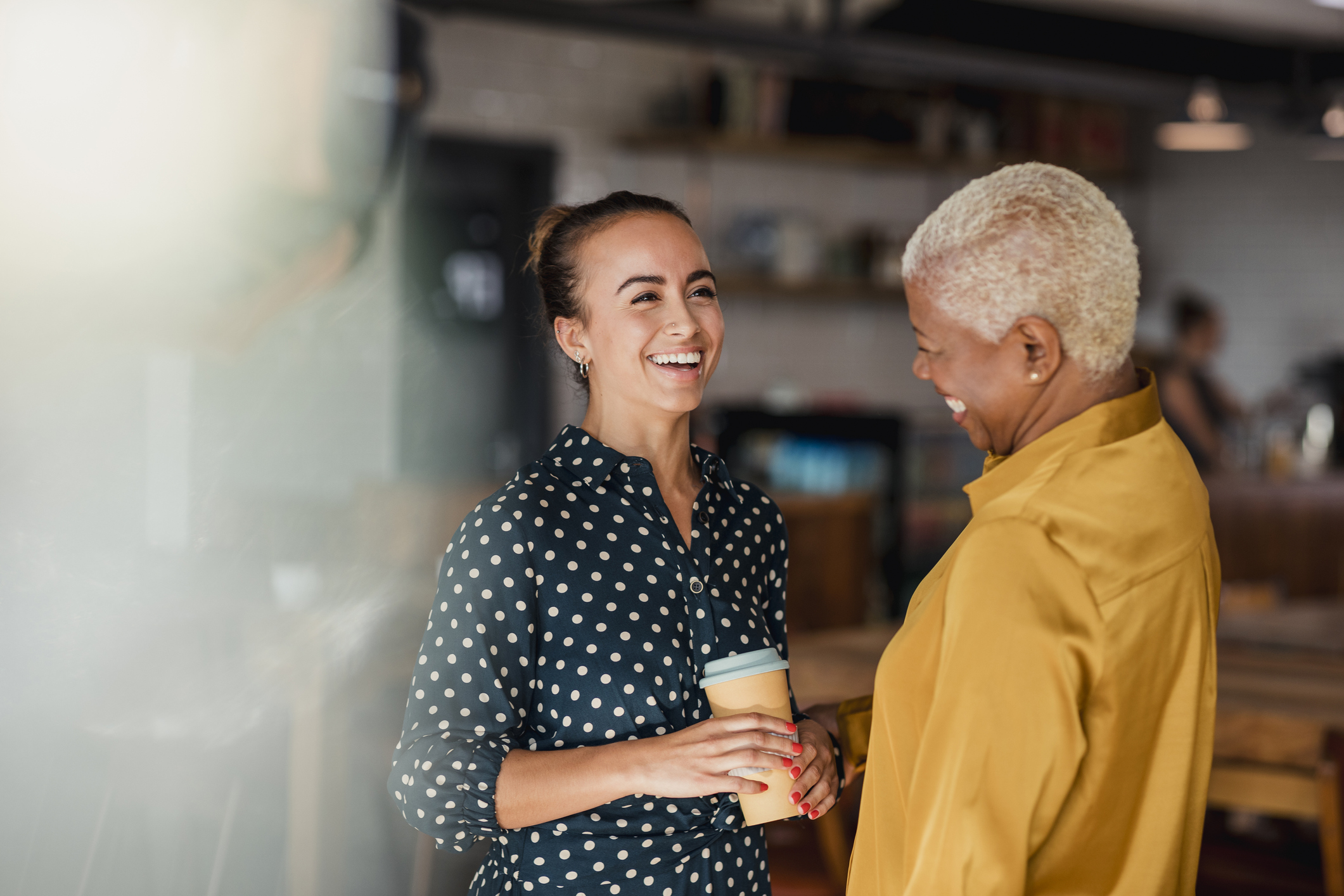 Two women colleagues laughing while standing in a cafe at their workplace. One of the women is holding a take out hot drink cup